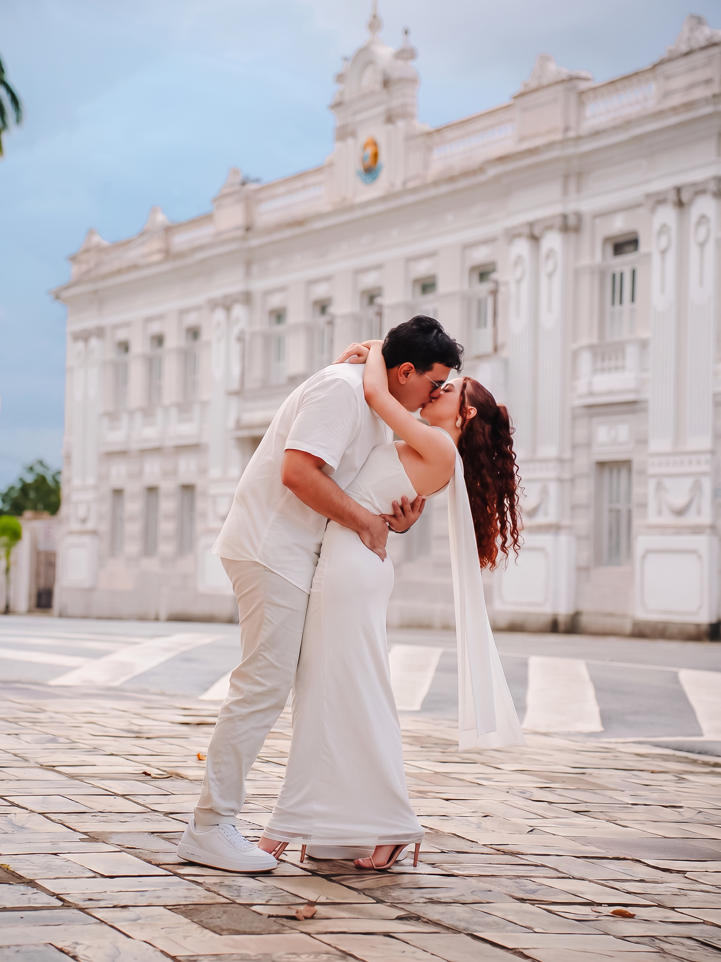 Maria e Sérgio se beijam diante de uma fachada branca, vestidos em tons claros.